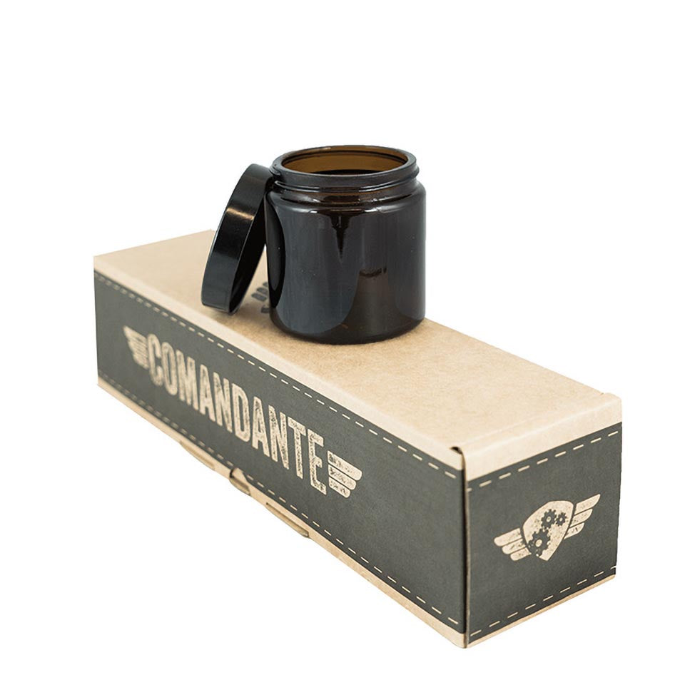 En brun glaskrukke fra Comandantes 4-pack sidder på sin mærkevare beige og sorte æske med et gear og vinger-logo. Det lågløse kaffebønneglas afslører æskens syede designaccenter.