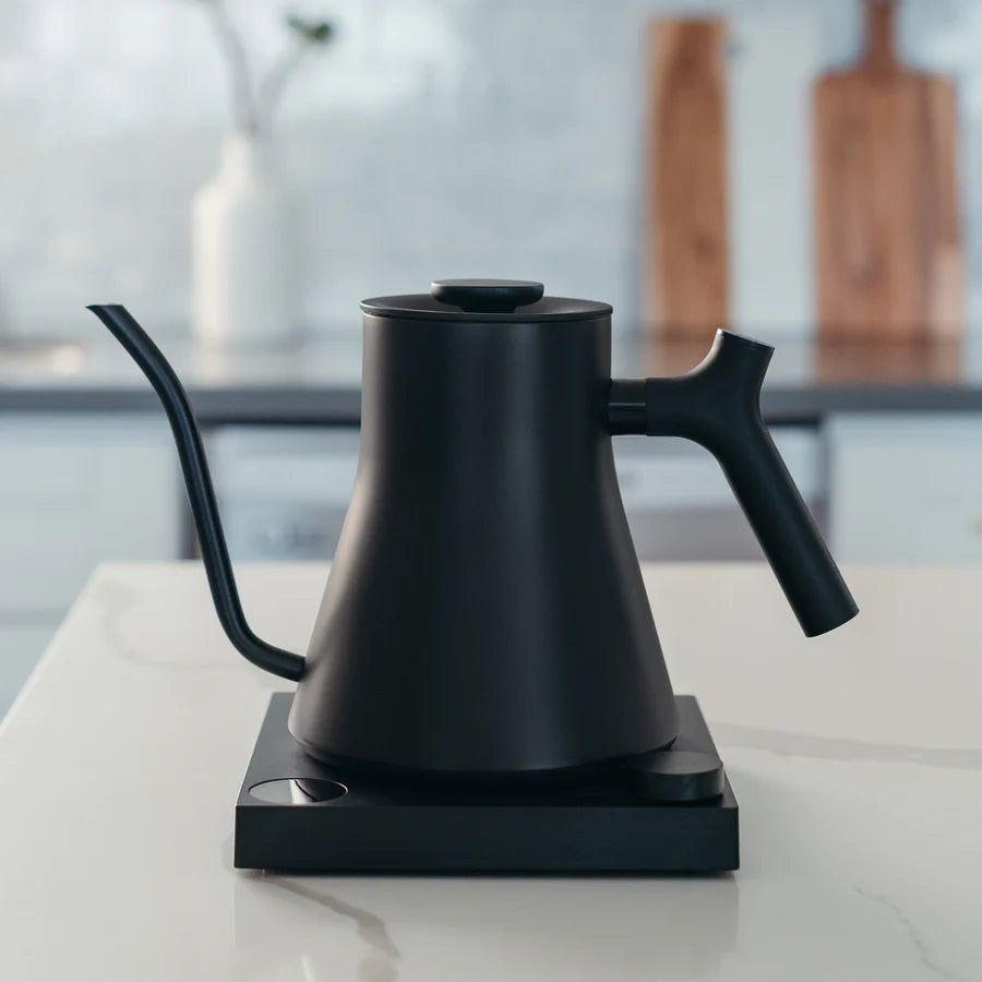 En elegant Fellow Stagg EKG PRO-kedel i sort med præcis temperaturkontrol sidder på en moderne bordplade. Den slørede baggrund afslører to træplader og en hvid vase, der fremhæver kedlens minimalistiske design og glatte finish for en moderne æstetik.