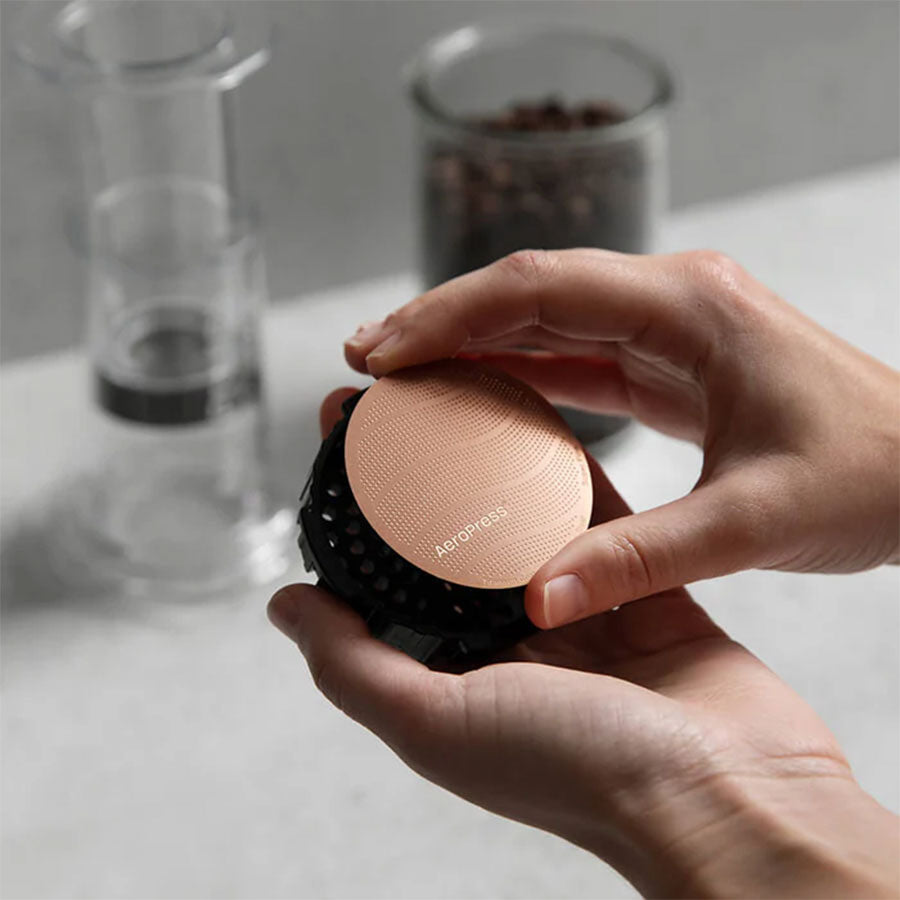 En persons hænder holder et AeroPress Gold Tone-filter fra Aeropress, fremstillet af 316 rustfrit stål, med sløret kaffeudstyr og bønner i baggrunden. Dette genanvendelige metalfilter er både stilfuldt og praktisk.