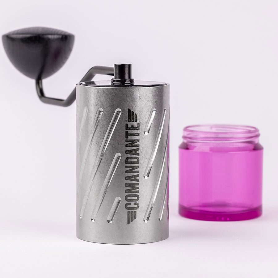 En Comandante C60 Baracuda - Stone Wash manuel kaffekværn med et sort håndtag står ved siden af en tom, gennemsigtig lyserød beholder til kaffegrums på en hvid baggrund.