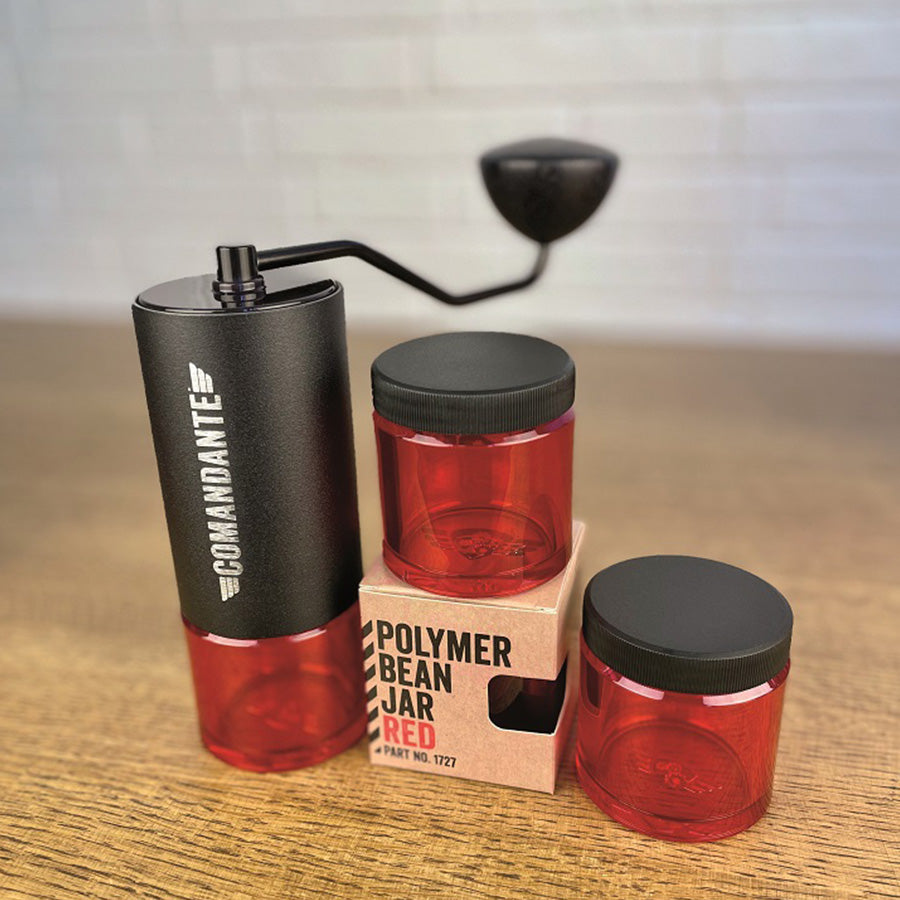 En sort manuel kaffekværn med et håndtag sidder ved siden af tre røde Comandante Polymer Bean Krukker, hvilket forbedrer din kaffeløsning. To krukker er stablet på en pap "Polymer Bean Jar Red" boks, alle vist på en træoverflade mod en hvid murstensvæg som baggrund.