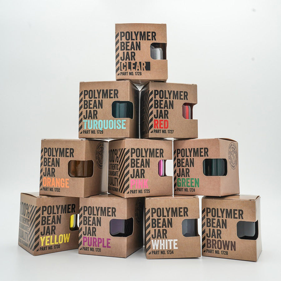 En pyramide af Comandante papkasser mærket "Polymer Bean Jar med låg, Rød" i forskellige farver inklusive klar, rød, turkis og mere. Hver æske har en udskæring med farverige bønner, perfekt til din kaffeløsning med Comandante C40.