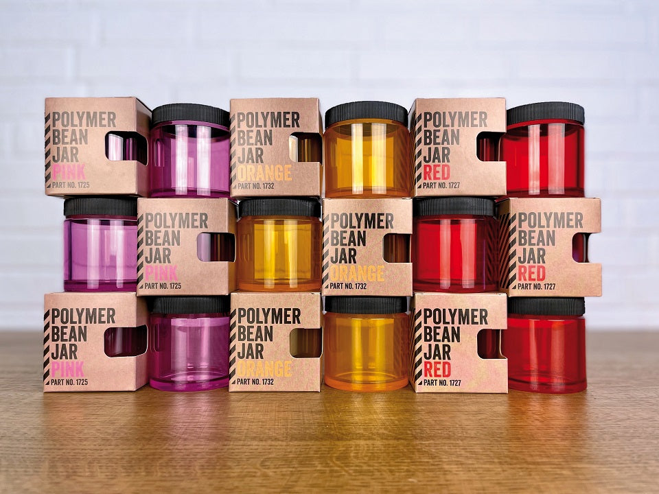 En stak af Comandante Polymer bønnekrukker med låg - Comandante C40 kompatible - i lilla, orange og rød er indkapslet i udskårne papkasser, der viser deres nuancer, arrangeret på en træoverflade mod en hvid murstensbaggrund.