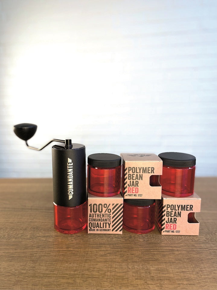 En Comandante manuel kaffekværn med sort håndtag sidder ved siden af "Comandante Polymer Bean Jar med låg, Rød" beholdere og viser din ideelle kaffeløsning. Nogle krukker er stablet i brune kasser på en træoverflade mod en neutral baggrund.