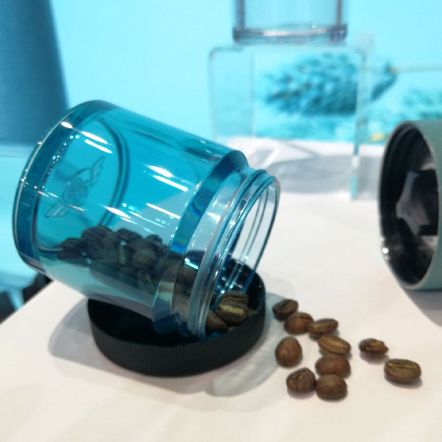 En gennemsigtig "Comandante, Polymer Bean Jar med låg, Tyrkis" vælter og spilder kaffebønner på en hvid overflade. Låget er i nærheden, da baggrunden sløres med antydninger af blåt og sølv, der inkarnerer fødevaresikker-standarder for din ro i sindet.