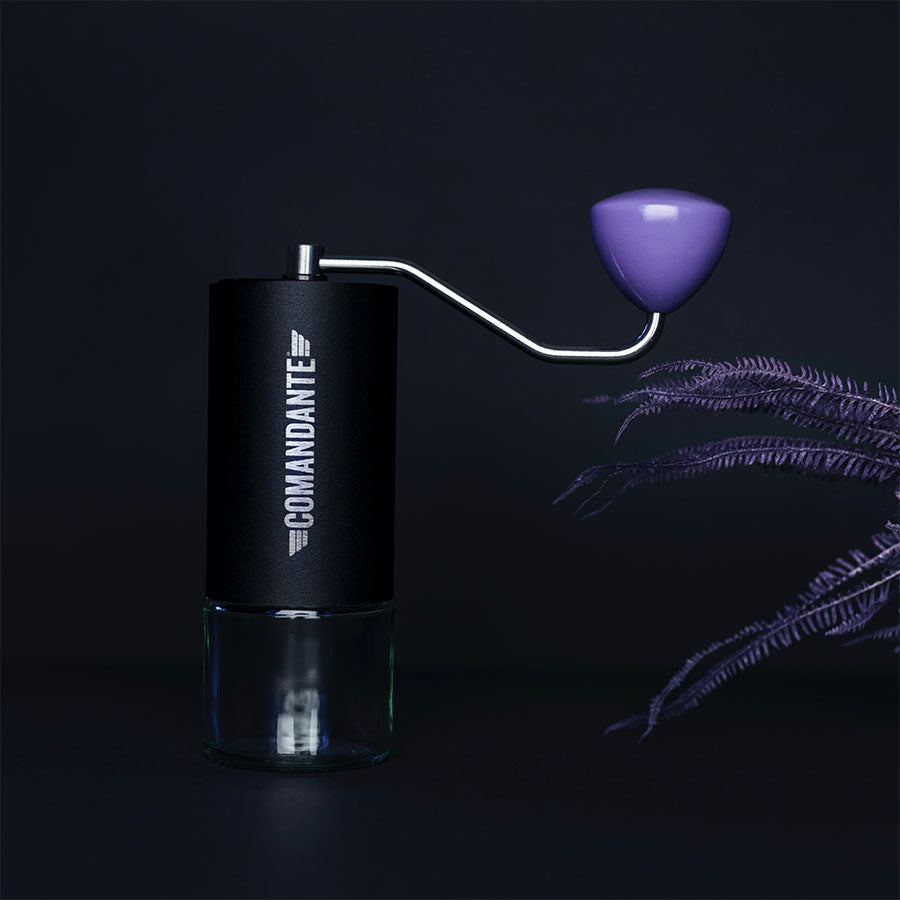 Mod en mørk baggrund med lilla bregne-lignende blade har den slanke Comandante kaffekværn en glasbund og metalhåndtag, dækket af en elegant ø 44 mm bøgetræ Lilla træknop.