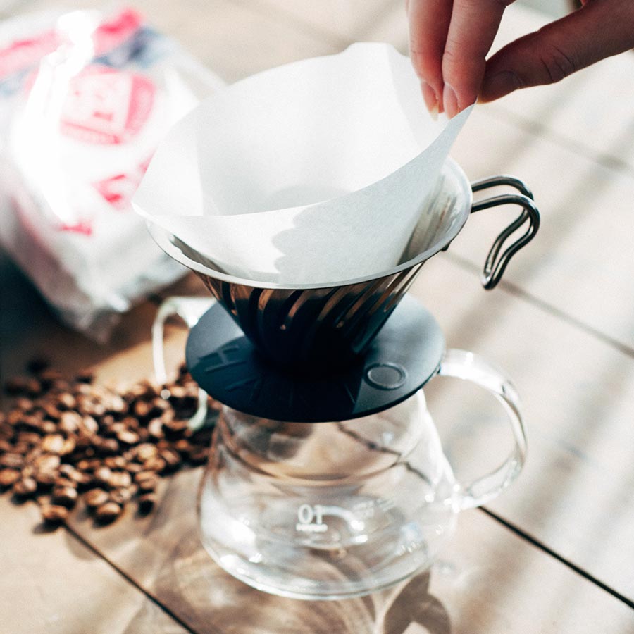 En hånd sætter en Hario, Papir Filter, Størrelse 01 i en metal hælde-over drypper på en glaskaraffel. Kaffebønner ligger på træoverfladen og booster kaffebrygningsoplevelsen. En sløret kaffepose sidder i baggrunden, mens naturligt sollys blødt oplyser scenen.