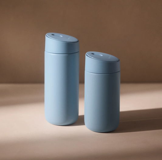 To matte lyseblå Fellow Carter Slide Mug Termo Krus fra Fellow står side om side på en svagt oplyst flade, den ene højere end den anden. Disse slanke, minimalistiske varmebevarende rejsekrus står på en neutral brun baggrund.