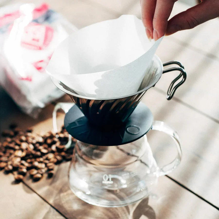 En hånd placerer et papirfilter i en kaffedrypper af metal, der står på en glaskaraffel, mens kaffebønner og en pose kaffe er synlige på en træplade i nærheden.
