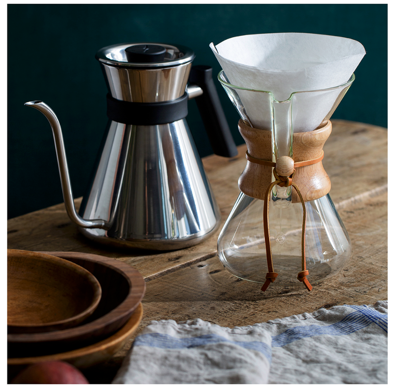 En svanehals-kedel i metal og en Chemex-kaffemaskine i glas med trækrave og papirfilter står på et træbord ved siden af træskåle og en klud.
