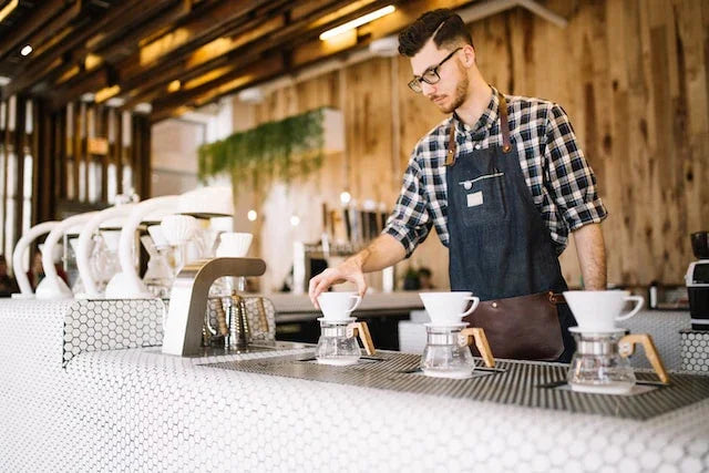 En barista iført briller, ternet skjorte og forklæde tilbereder kaffe ved hjælp af pour-over-dryppere på en moderne kaffebar med trævægge og en elegant disk.