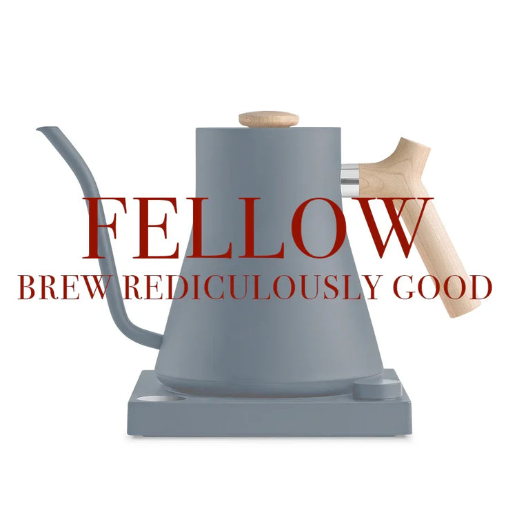 En matgrå elkedel med træhåndtag og -knap på en matchende base. Overlejret rød tekst lyder: "KOLLEGA BRYGGER FANTASTISK GODT.