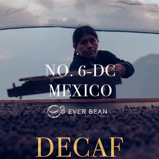 En person i mørkt tøj bearbejder kaffebønner udendørs under en baldakin. Teksten lyder: "No. 06-dc - Mexico Decaf, Dark Roast (Øko), EverBean." De landlige bjerge antyder oprindelsen af denne økologiske koffeinfri espresso.