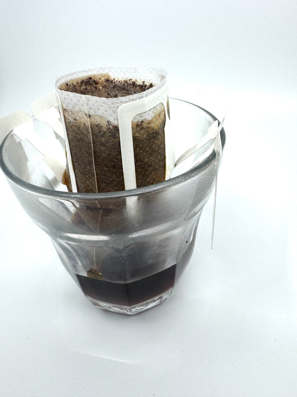 Et No. 07 - Tanzania, Single Serve drypkaffefilter fra EverBean ApS. er fastgjort til en glaskop, der indeholder våd Vasket Arabica-grums, mens den bryggede kaffe samler sig i bunden, mod en almindelig hvid baggrund.