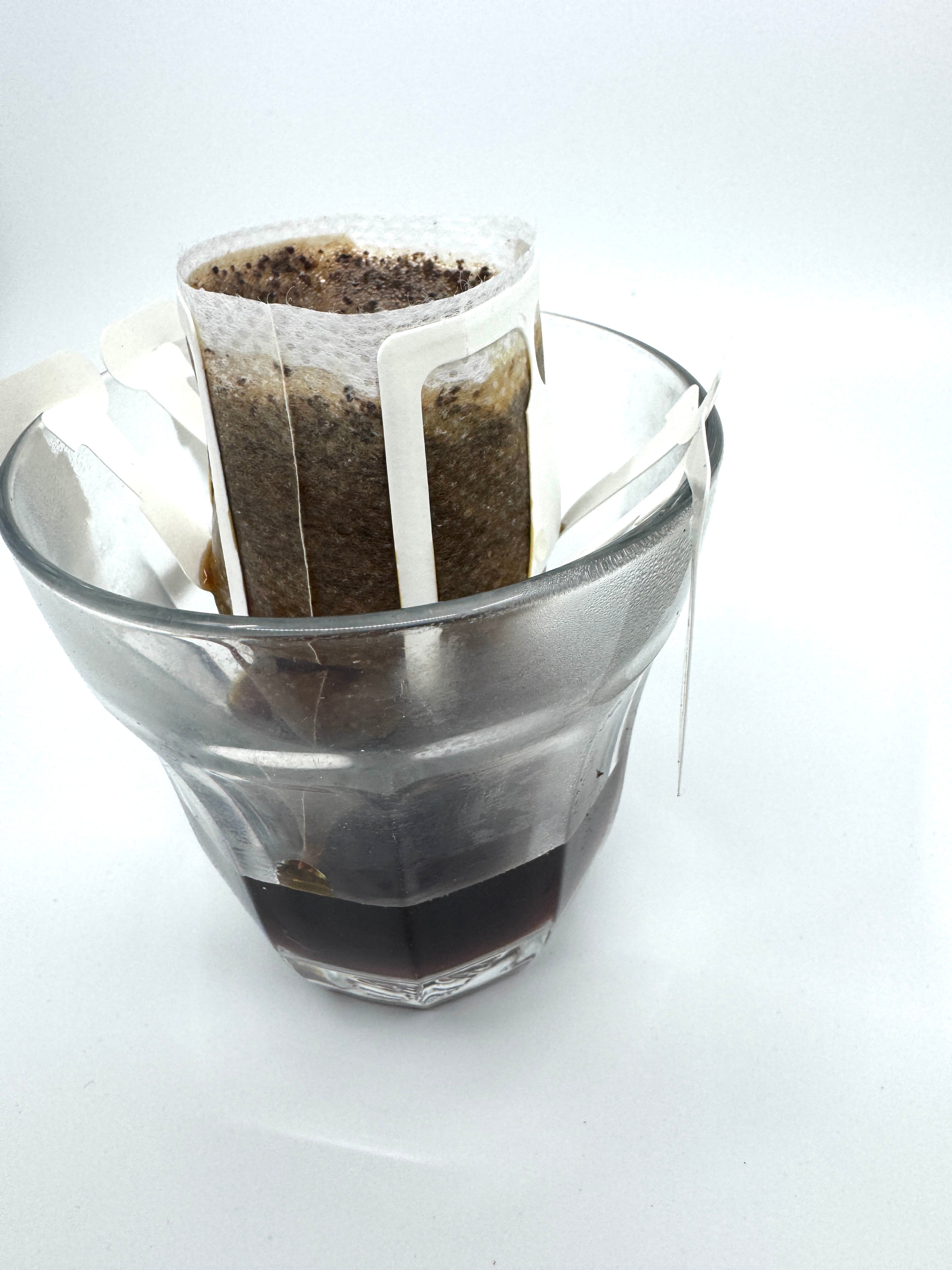 Et No. 07 - Tanzania, Single Serve drypkaffefilter fra EverBean ApS. er fastgjort til en glaskop, der indeholder våd Vasket Arabica-grums, mens den bryggede kaffe samler sig i bunden, mod en almindelig hvid baggrund.