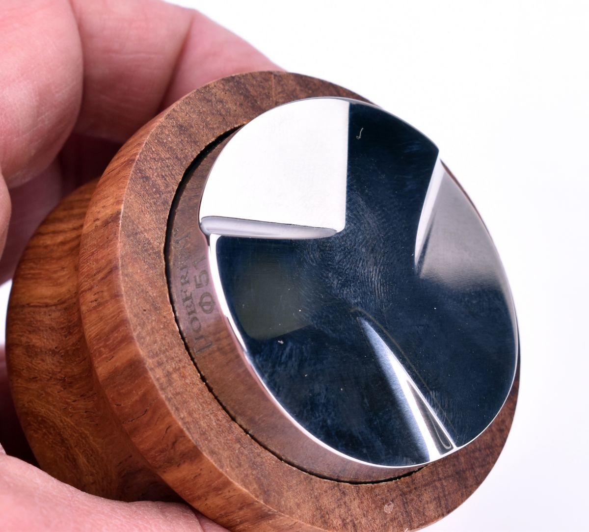En hånd holder et skinnende Joe Frex Coffee Level Tool med en 58 mm reflekterende metaloverflade og en poleret Palisander trækant, med et rigt åremønster og glat, buet design.