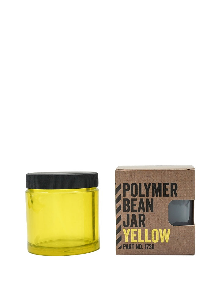 En gennemsigtig gul Comandante polymer krukke mærket "Polymer Bean Jar med låg, Gul" med sort låg er ved siden af en brun æske. Den fede tekst på æsken lyder "POLYMERBØNNEKRUDE GUL DEL NR. 1730", hvilket sikrer, at det er fødevaresikker til sikker opbevaring.