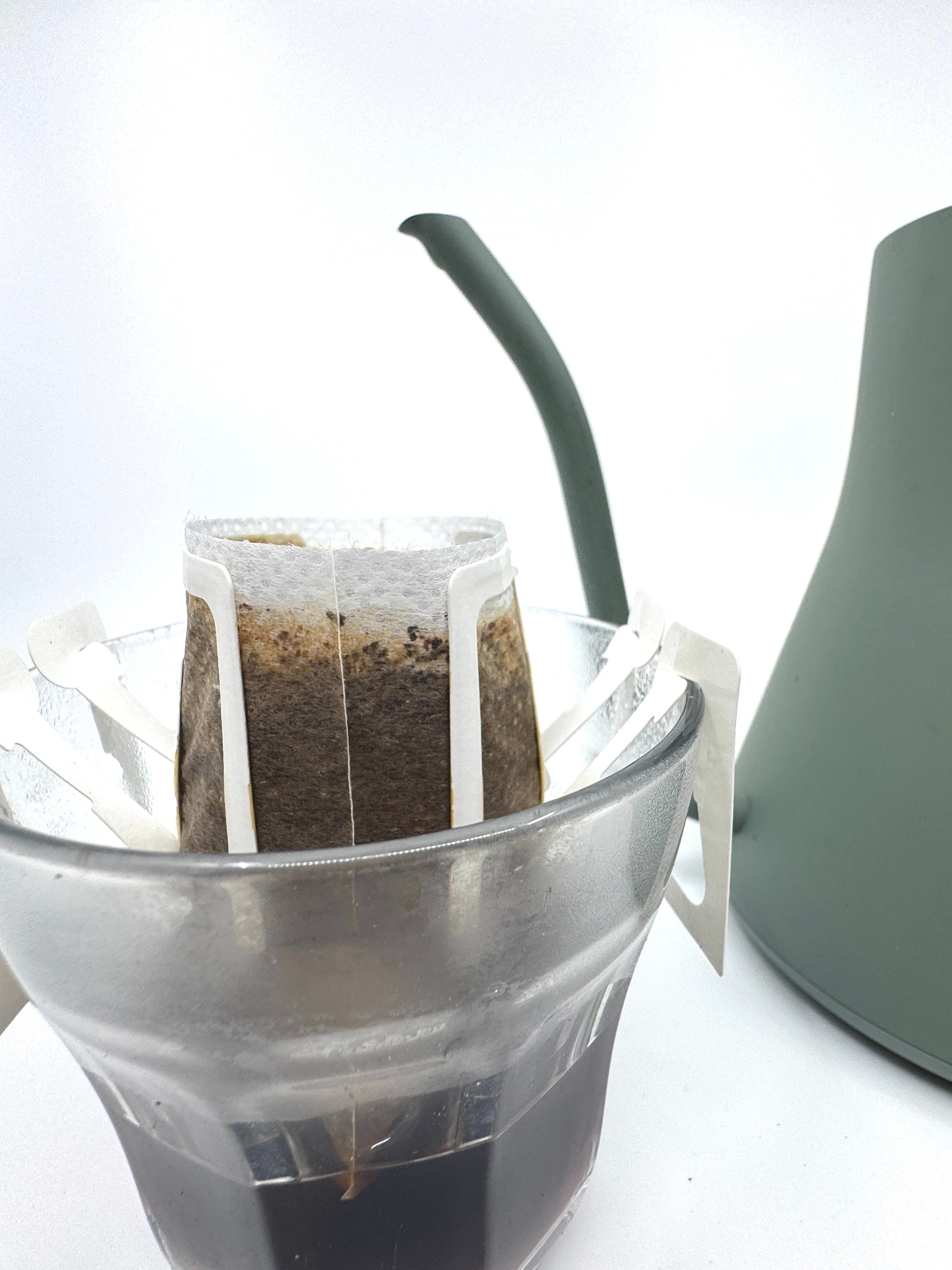 Et No. 07 - Tanzania, Single Serve pour-over kaffefilter fra EverBean ApS. er sat på et glaskrus, mens der hældes varmt vand fra en grøn svanehals-kedel i baggrunden mod en hvid overflade.