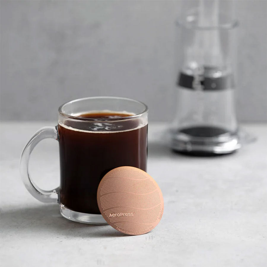 Et klart glaskrus med sort kaffe står på en grå overflade ved siden af det runde AeroPress Gold Tone-filter fra Aeropress, med en sløret AeroPress-kaffemaskine i baggrunden, der antyder førsteklasses bryggekvalitet.