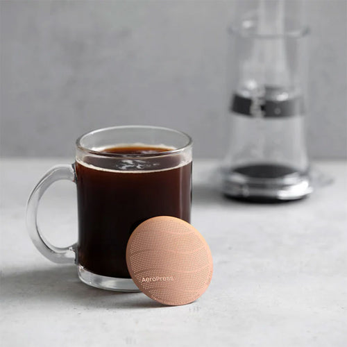 Et klart glaskrus med sort kaffe står på en grå overflade ved siden af det runde AeroPress Gold Tone-filter fra Aeropress, med en sløret AeroPress-kaffemaskine i baggrunden, der antyder førsteklasses bryggekvalitet.