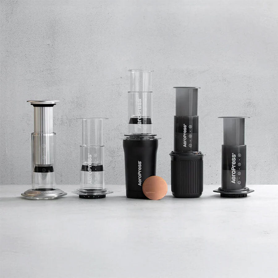 Fire Aeropress-kaffemaskiner er arrangeret på en grå flade, så man kan se deres klare og sorte dele ved siden af Aeropress Gold Tone-filteret fra Aeropress, der er fremstillet af holdbart, genanvendeligt 316 rustfrit stål.