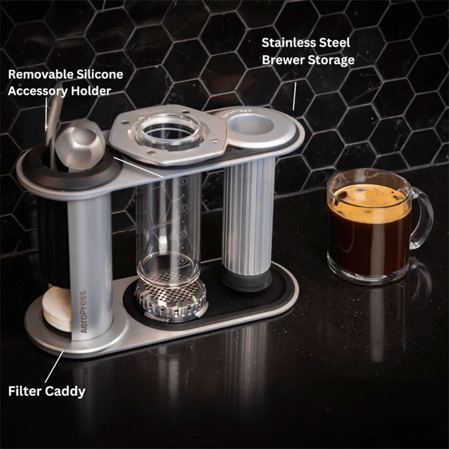 Aeropress Premium Organizer fra Aeropress har en aftagelig holder til silikonetilbehør, filterholder og opbevaringsplads til bryggeren i rustfrit stål. Ved siden af den står et glaskrus med kaffe på en sort bordplade med sekskantede fliser.
