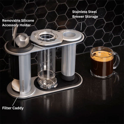 Aeropress Premium Organizer fra Aeropress har en aftagelig holder til silikonetilbehør, filterholder og opbevaringsplads til bryggeren i rustfrit stål. Ved siden af den står et glaskrus med kaffe på en sort bordplade med sekskantede fliser.