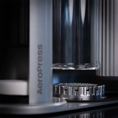 Nærbillede af Aeropress Premium Organizer fra Aeropress, der fremviser dens minimalistiske design og cylindriske kammer over et filter i rustfrit stål, med Aeropress-logoet synligt. Billedet fremhæver bryggemaskinens elegante komponenter.