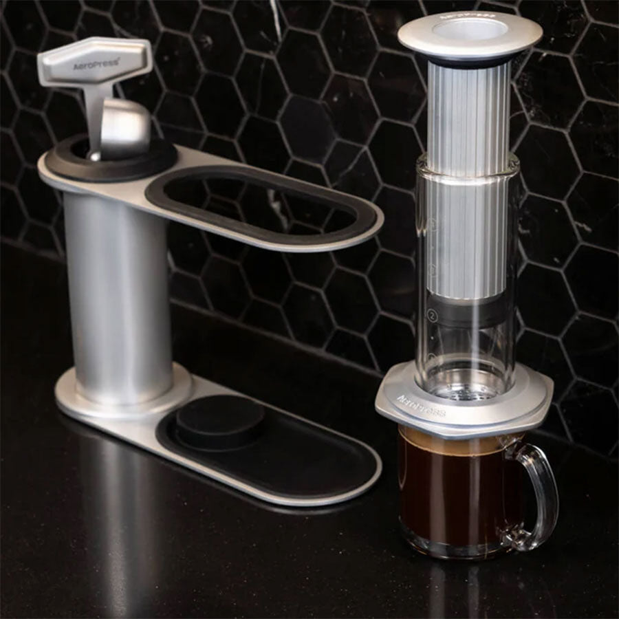 To Aeropress-kaffemaskiner står på en elegant sort bordplade med sekskantede fliser; den ene ved siden af et krus, den anden i brug – og fremviser den stilfulde Aeropress Premium Organizer fra Aeropress, perfekt til minimalistiske køkkener.