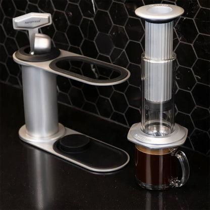 To Aeropress-kaffemaskiner står på en elegant sort bordplade med sekskantede fliser; den ene ved siden af et krus, den anden i brug – og fremviser den stilfulde Aeropress Premium Organizer fra Aeropress, perfekt til minimalistiske køkkener.