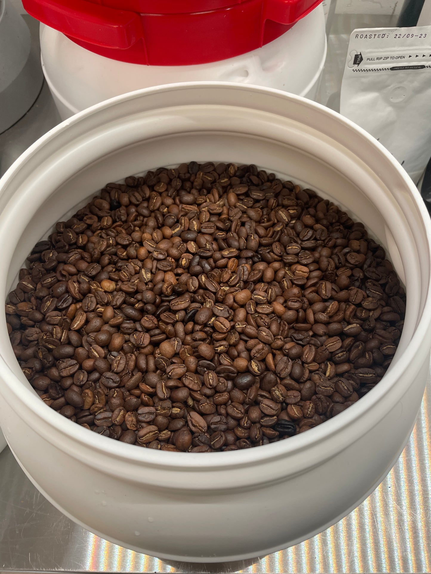 En hvid beholder fyldt med ristede Arabica bønner sidder på en reflekterende overflade. Den afslører mørkebrune bønner indeni, med et rødt og hvidt låg i nærheden. I baggrunden ses en mærket pose EverBeans No. 02 · Brasilen kaffe.
