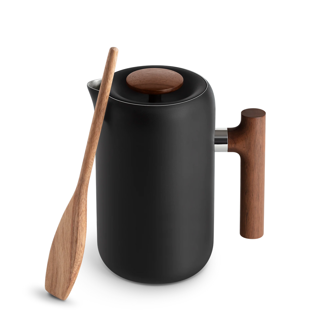Den slanke sorte Fellow Clara French Press, Valnød har vakuumisolerede vægge for optimal varmetilbageholdelse. Dets moderne design er fremhævet af et træhåndtag og låg, med en træspatel lænet mod det på en almindelig baggrund.