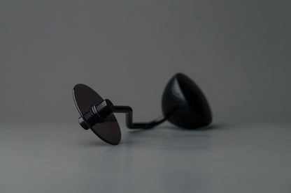 Et par sorte solbriller med ovale glas hviler på en grå overflade. Den ene linse vender opad, og armen ligner et Comandante® Sort Håndsving kværnhåndtag, hvilket skaber en minimalistisk og moderne stemning mod den glatte neutrale grå baggrund.