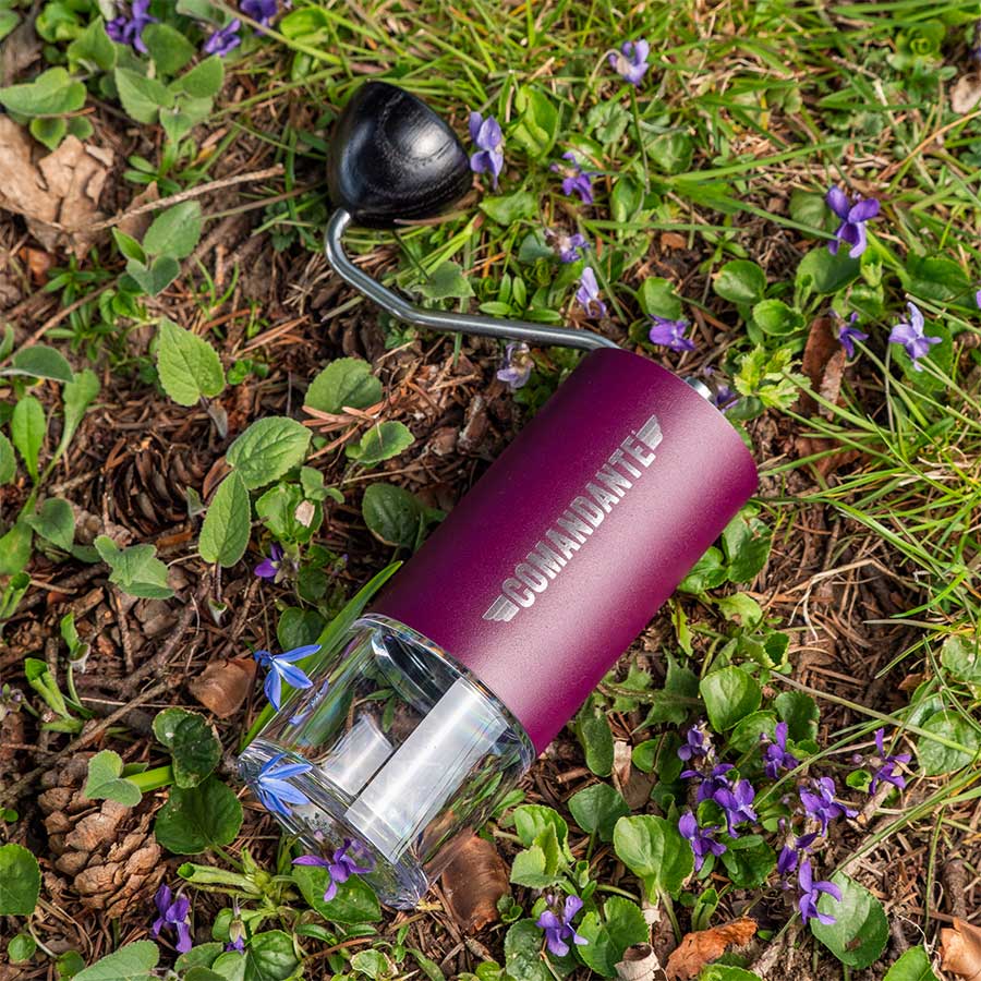 Et Comandante C40 MK4 Nitro Blade kaffekværn i Deep Purple fra Comandante med glasbund ligger på grønt græs og små lilla blomster udendørs.