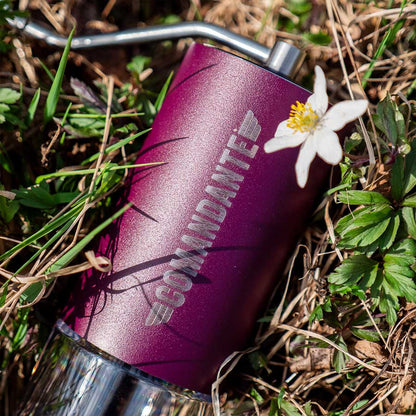 En Comandante C40 MK4 Nitro Blade, Deep Purple kaffekværn hviler på græs og små planter med en hvid blomst ved siden af.