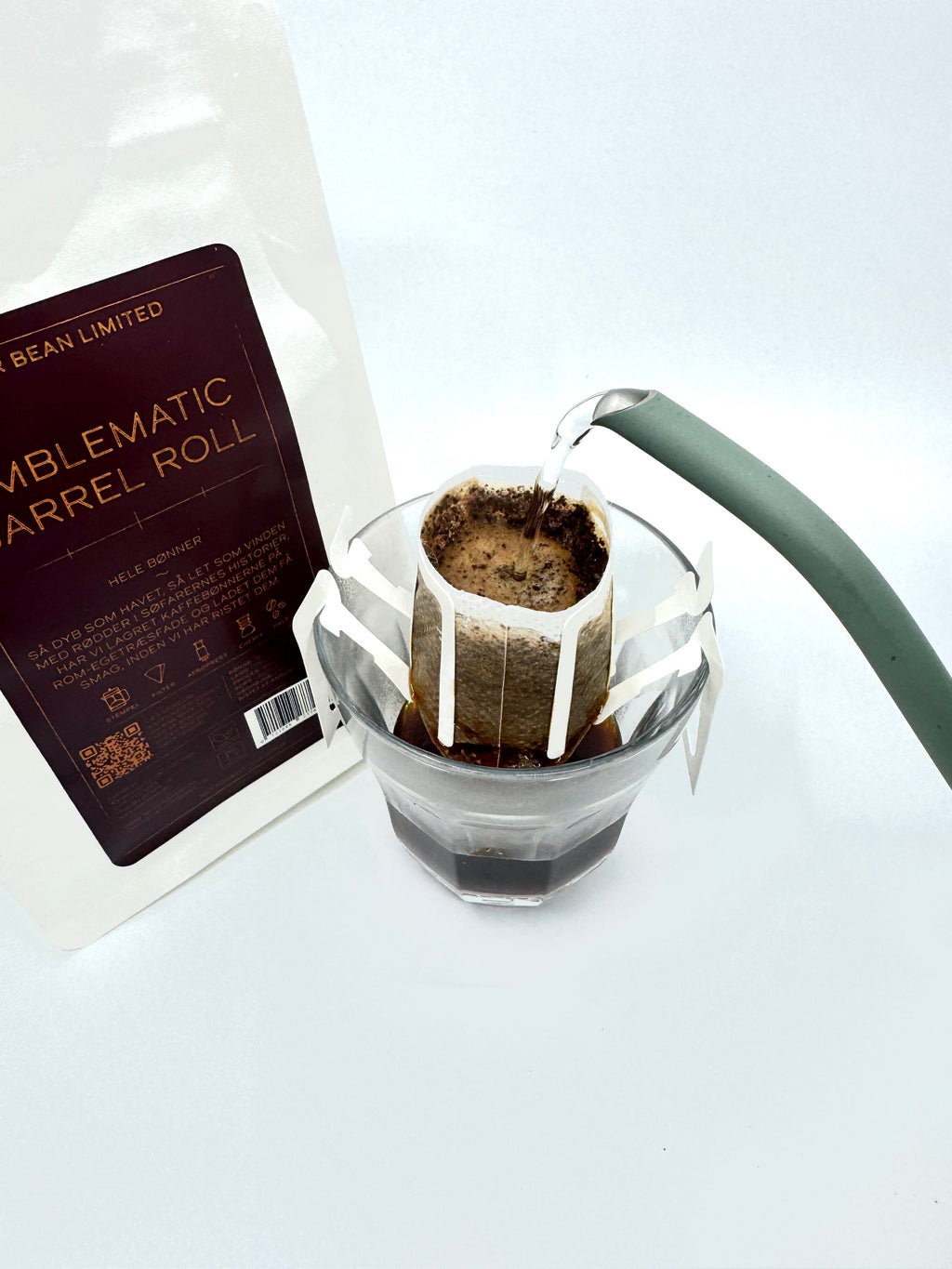 Nærbillede af en pour-over med varmt vand fra en grøn kedel på colombianske bønner i et papirfilter over glas ved siden af en hvid pose mærket "Emblematic Barrel Roll, Limited, Single Serve" fra EverBean Limited.