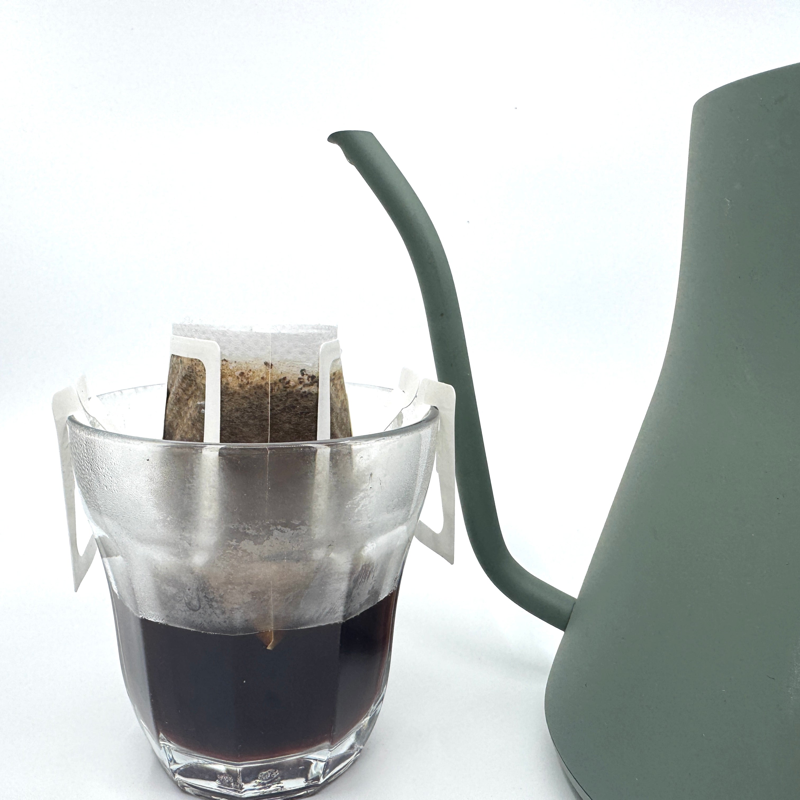 Et glas med en Single Serve, colombiansk maceration-kaffefilterpose fra EverBean Limited fastgjort til kanten, fyldt med brygget kaffe. Ved siden af står en delvis synlig grøn svanehals-kedel på en almindelig hvid baggrund.
