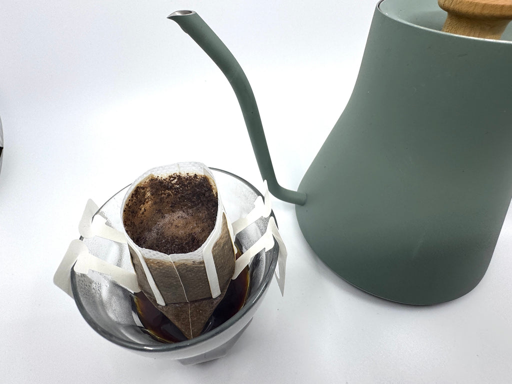 Nr. 03 · Burundi · Enkeltdosis-filterpose fra EverBean ApS. står på et glaskrus og fremhæves for sin lyse ristning, mens en grøn kedel med træhåndtag står ved siden af, klar til at brygge den perfekte kop kaffe.