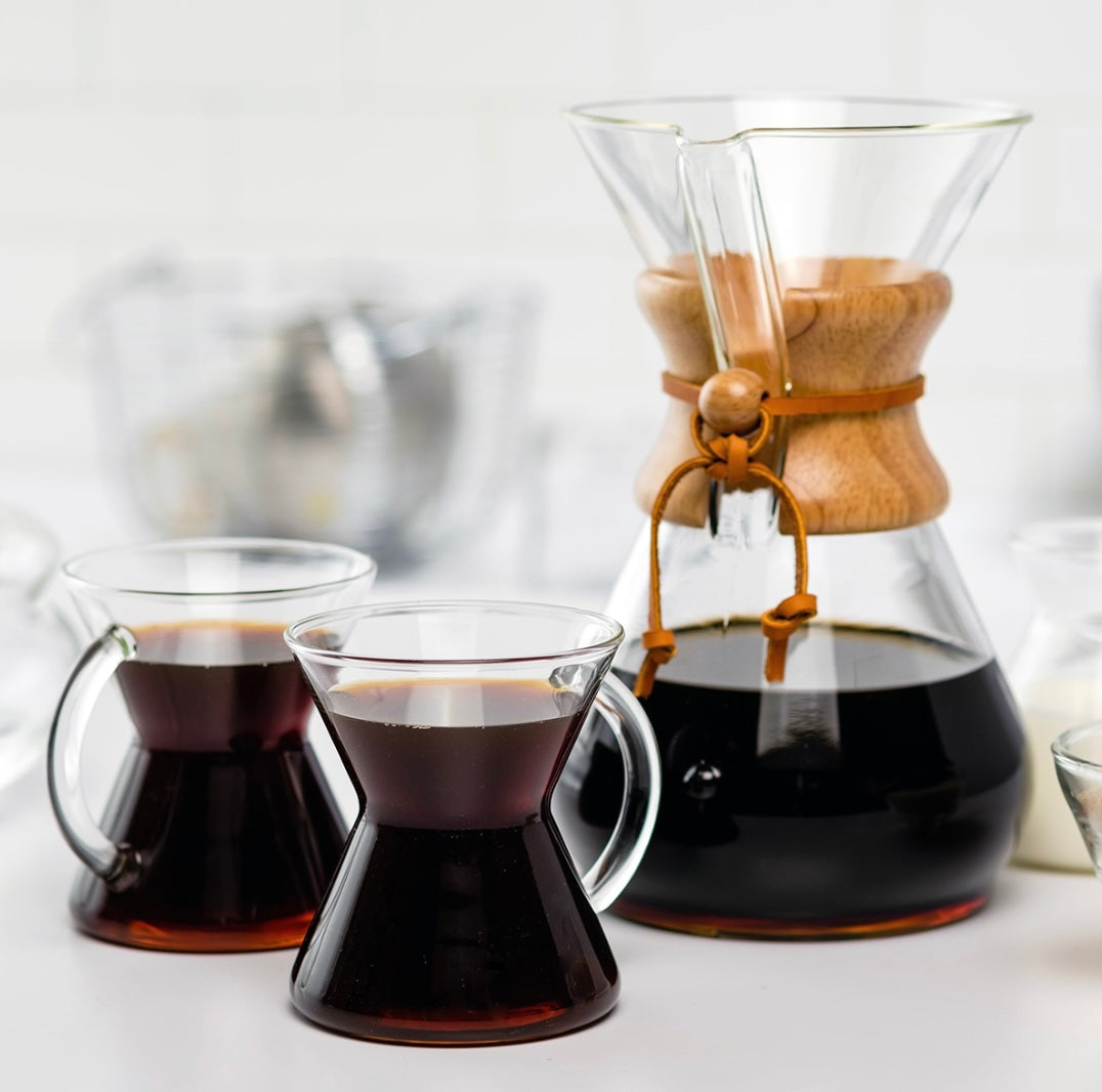 A Chemex Classic Kaffebrygger 8-kopper (CM-8A) fra Chemex står på en hvid overflade med sort filterkaffe, flankeret af to glas kopper friskbrygget kaffe. I baggrunden anes blødt slørede køkkenartikler.