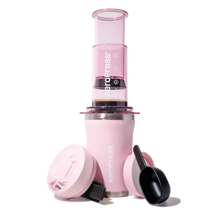 En AeroPress Go Plus fra Aeropress i pink, vist med stemplet sat i, matchende pink termokop, to pink låg, en sort scoop og en sort omrører arrangeret på en hvid baggrund.