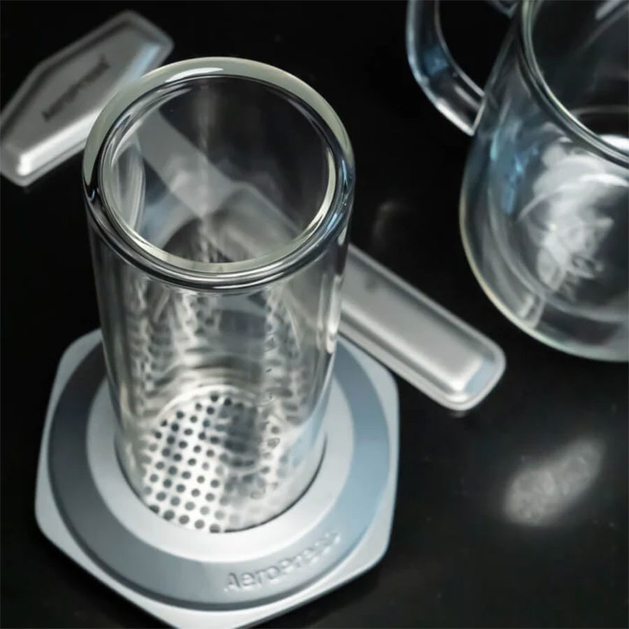 Et nærbillede af Aeropress, Premium kaffemaskine fra Aeropress på en sort overflade med filterhætten fjernet fremhæver den innovative 3-i-1 bryggeteknologi, mens et klart glaskrus er delvist synligt til højre.