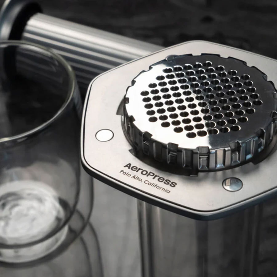 Nærbillede af Aeropress Premium kaffemaskine fra Aeropress med metalfilter og 3-i-1-brygningsteknologi. Aeropress-logoet er synligt på sølvkanten ved siden af en klar glaskop på en mørk overflade.