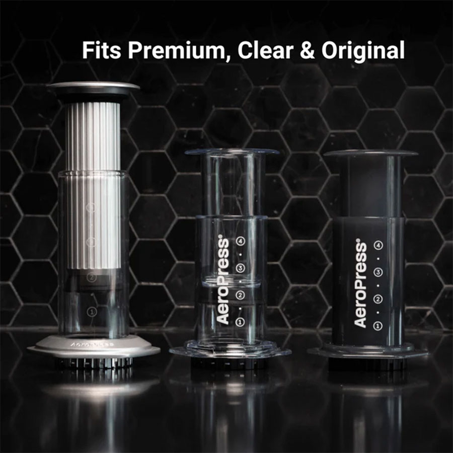 Kaffemaskinerne AeroPress Premium, Clear og Original fra Aeropress står side om side på sorte, sekskantede fliser med teksten "Fits Premium, Clear & Original" over dem, hvilket fremhæver deres 3-i-1-brygningsteknologi.