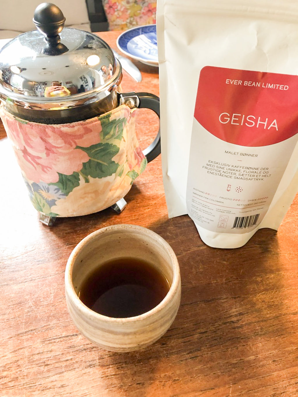 En kop sort kaffe står på et træbord ved siden af en blomstret French press og en hvid pose med EverBean Limiteds Limited, Columbia Geisha, der fremhæver en af de særlige kaffer for sande entusiaster.