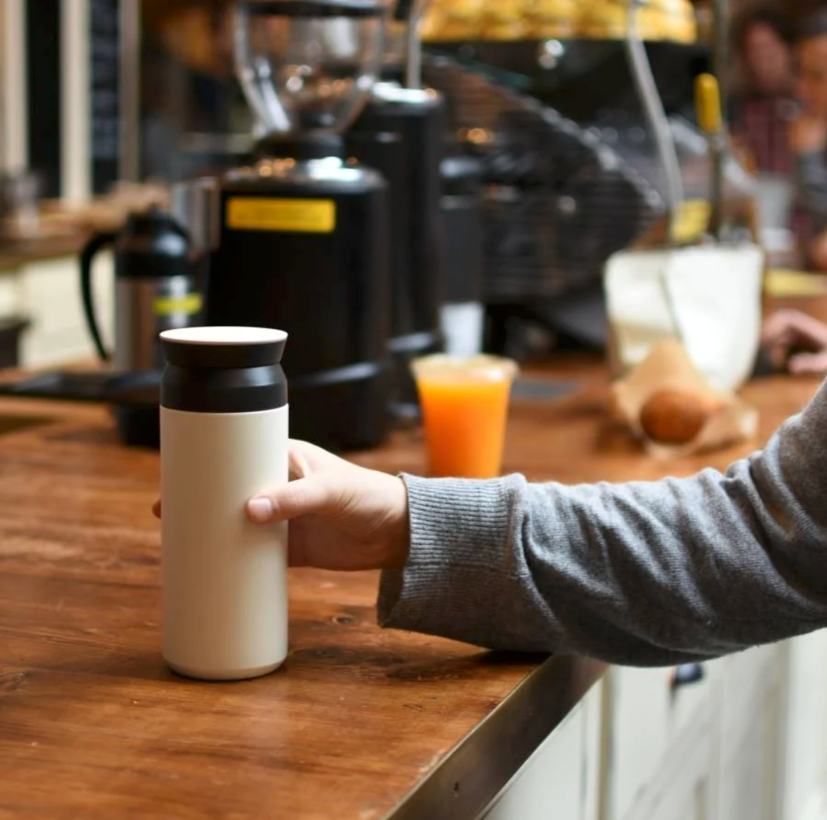 En person i en grå sweater holder en Kinto Travel Tumbler på en cafédisk i træ. Bag den er der et appelsinjuiceglas, en sort kaffekværn og kager, der alle understøtter deres bevidste og fleksible livsstil.