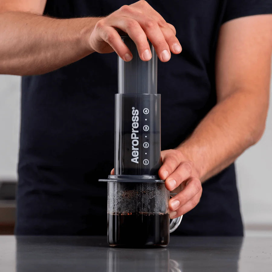 En person iført mørk skjorte trykker på en Aeropress Original kaffebrygger fra Aeropress, der er placeret over et krus med kaffe i klart glas. Kun deres overkrop og hænder er synlige.