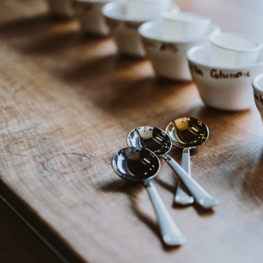 Tre metalskeer ligger på et træbord ved siden af hvide skåle med skrift på kanterne, mens en Brewista Professional Cupping Spoon (Titanium - Black) subtilt forstærker den bløde belysning og skaber en indbydende atmosfære perfekt til at nyde smag.