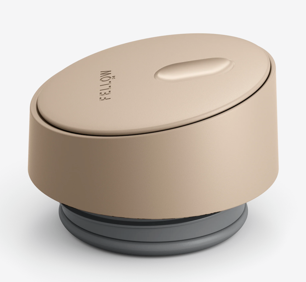 Et beige, glat, cylindrisk låg med "FELLOW" og en oval fordybning, der er designet til at passe til Fellow Carter Slide Mug Termo Krus, sidder på en grå base og giver et stilfuldt look.