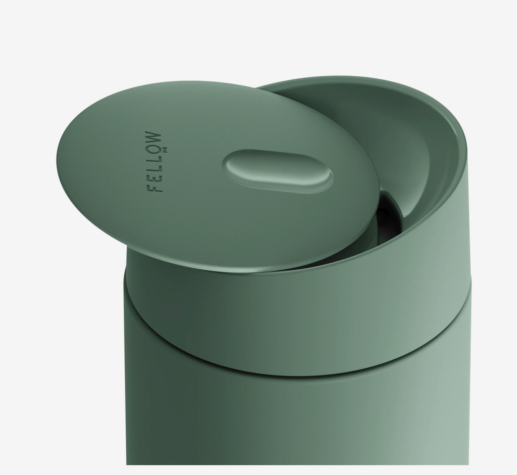 Et matgrønt Fellow Carter Slide Mug termo krus med delvist åbent låg og "FELLOW" trykt på toppen. Den keramiske belægning holder din drik frisk og smagfuld.