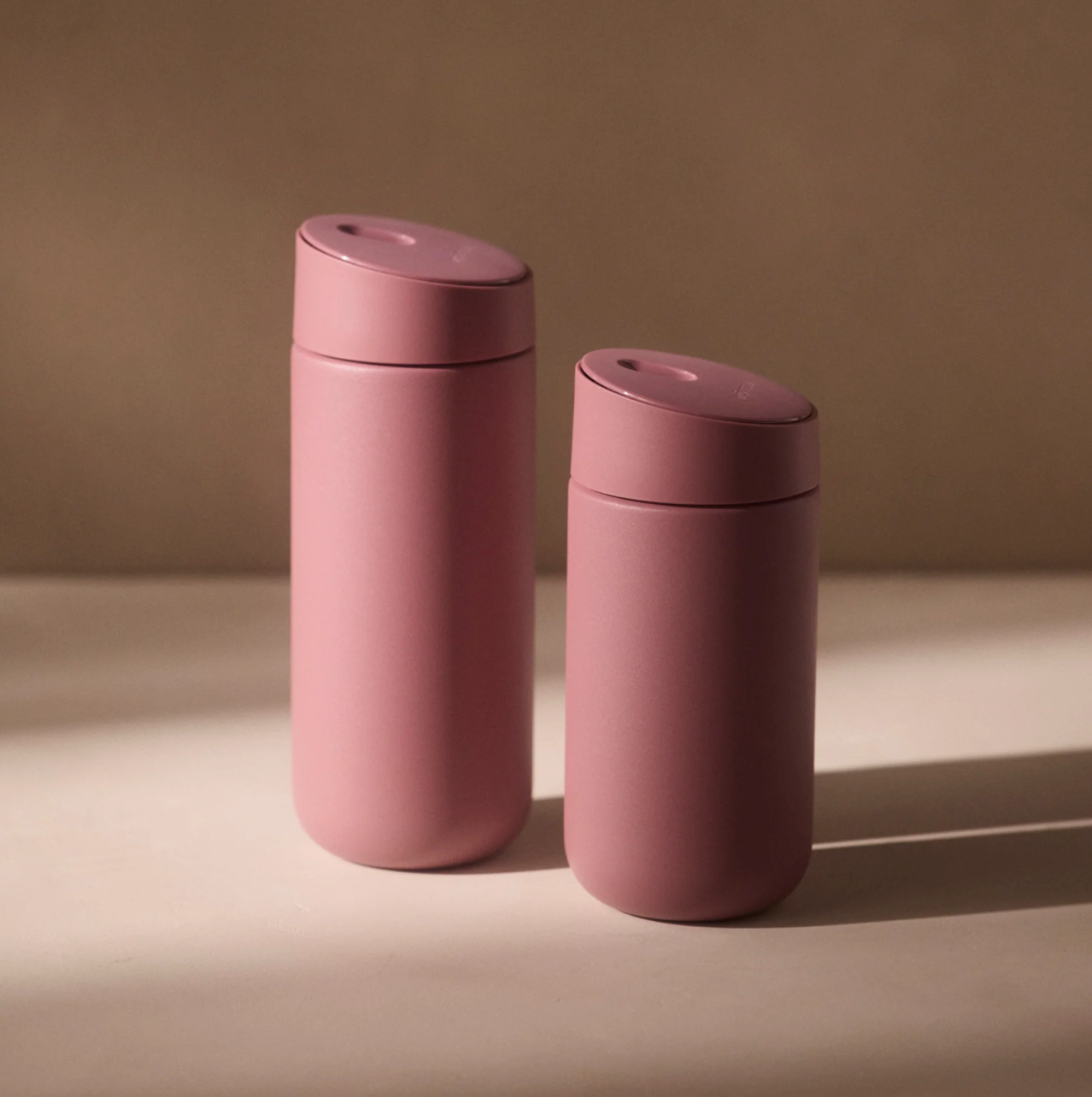 To Fellow Carter Slide Mug Termo Krus i mat rosa med afrundede låg står side om side på en beige overflade, blødt oplyst. Hvert Fellow-krus har keramisk belægning for varig kvalitet mod en neutral baggrund.